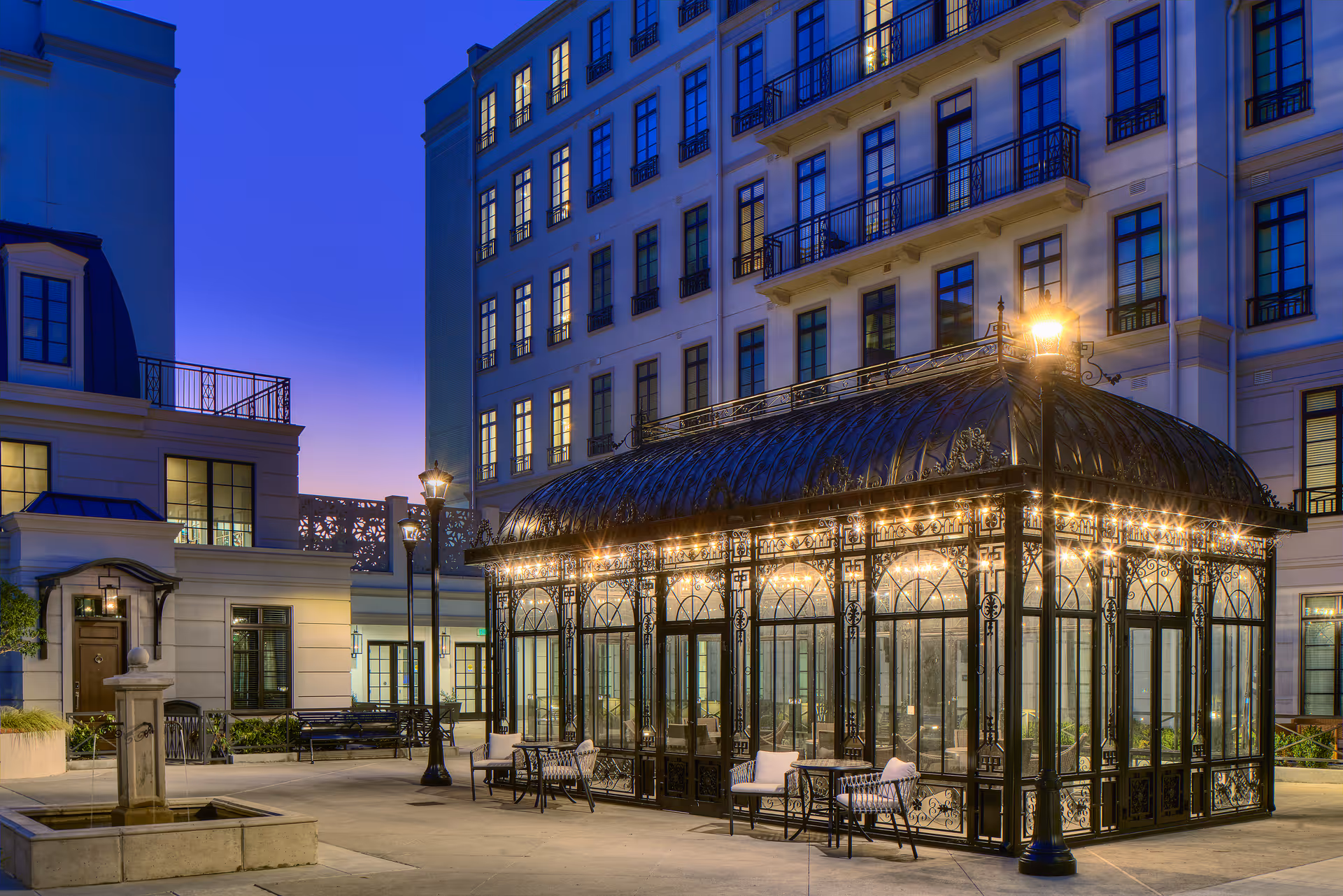 Outdoor courtyard area at dusk featuring a decorative glass and wrought iron pavilion with string lights, surrounded by multi-story residential buildings with lit windows. There are benches and a small fountain in the courtyard.