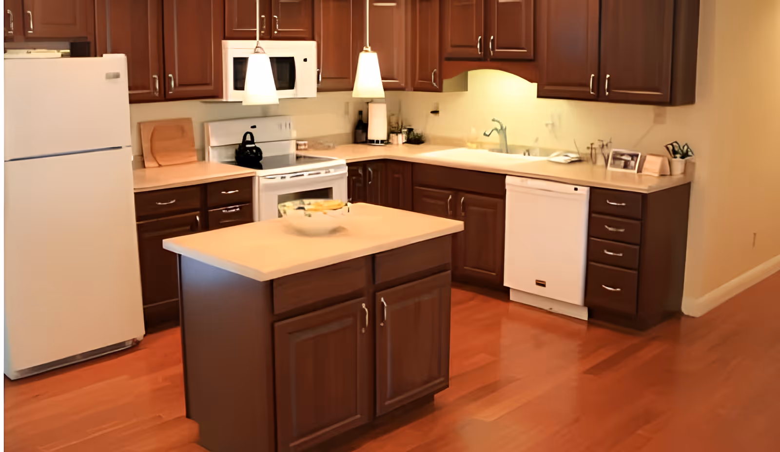 A kitchen with wooden cabinets, a white refrigerator, a white stove with a black kettle on it, a white microwave above the stove, a white dishwasher, and a small kitchen island with a bowl of bananas on top. The kitchen has hardwood flooring and two pendant lights hanging above the island.