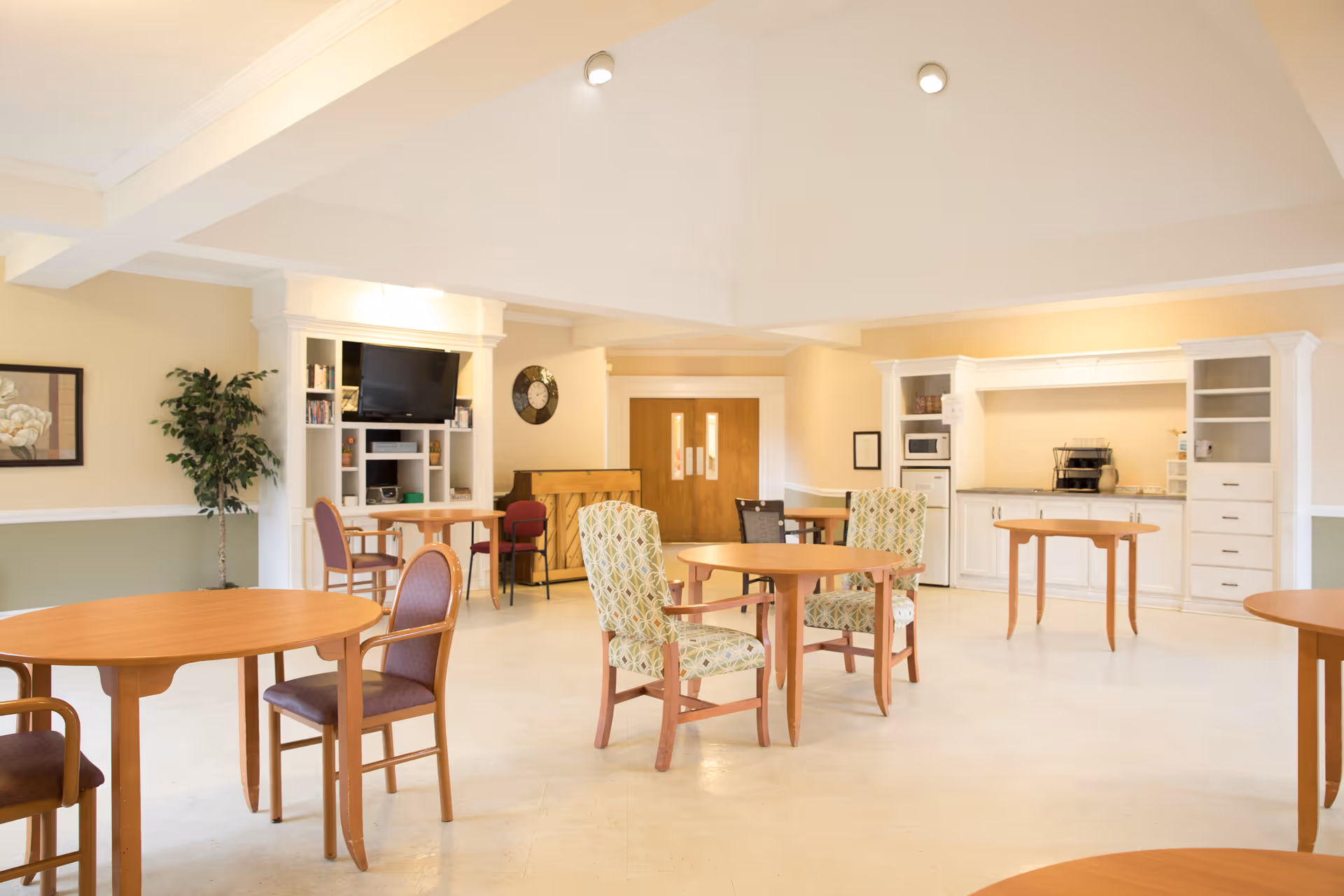 A bright and spacious common area in a senior living facility with multiple round wooden tables and chairs arranged throughout the room. The walls are painted in light colors with white trim, and there is a built-in entertainment center with a TV and shelves. A piano is positioned against the far wall near double wooden doors. The room also features a kitchenette area with white cabinets, a microwave, and a coffee maker.