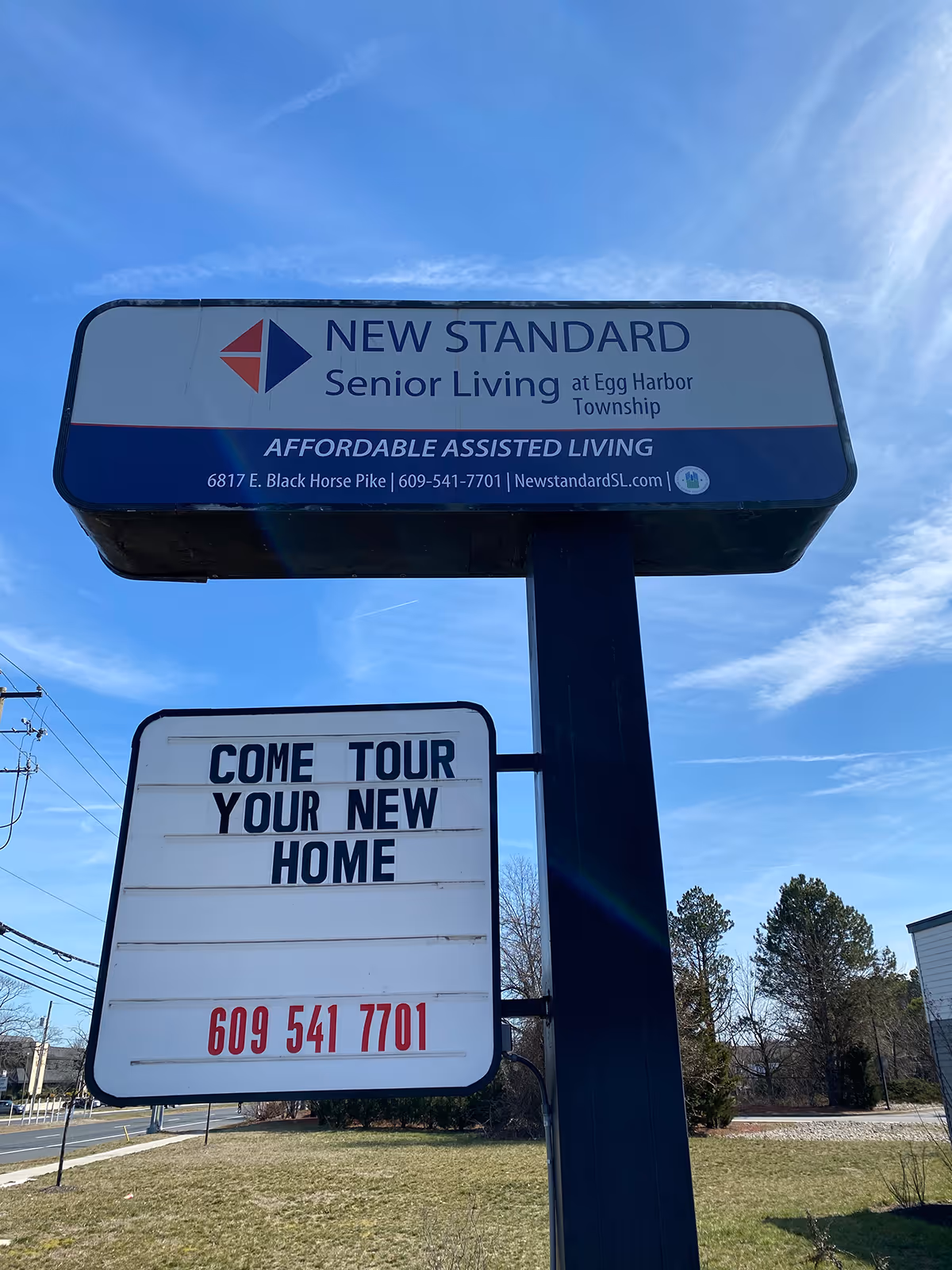 Outdoor sign for New Standard Senior Living at Egg Harbor Township with a message board below reading 'COME TOUR YOUR NEW HOME' and a phone number 609 541 7701, set against a blue sky and grassy area.