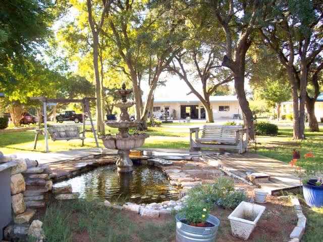 Courtyard with a central fountain and pond, wooden swings and benches under large trees in front of a single-story building.