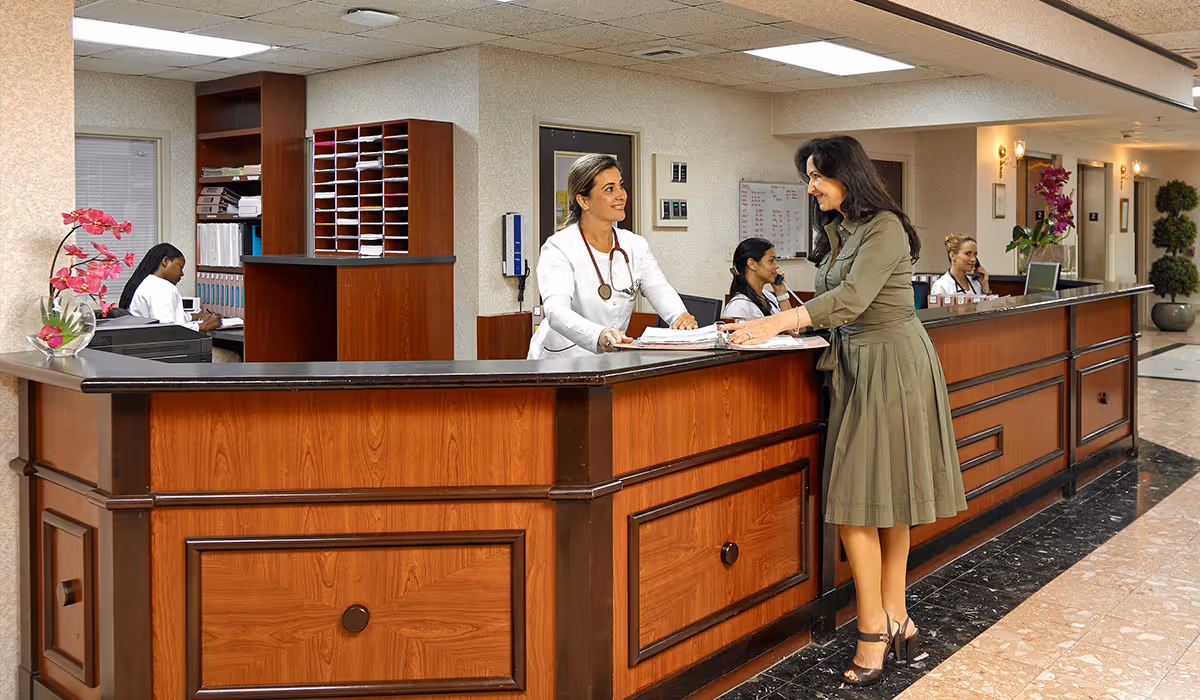Reception area in a nursing and rehabilitation center with a wooden front desk. A woman in a green dress is talking to a staff member wearing a white coat with a stethoscope. Two other staff members are seated behind the desk working. The area is well-lit with ceiling lights and decorated with pink orchids and potted plants.