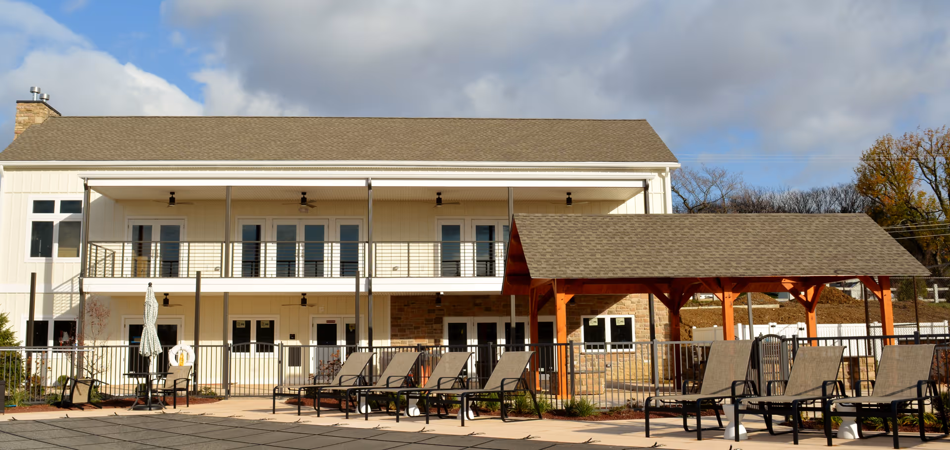 Outdoor view of a two-story senior living facility building with a covered patio area and multiple lounge chairs arranged around a fenced pool area under a partly cloudy sky.