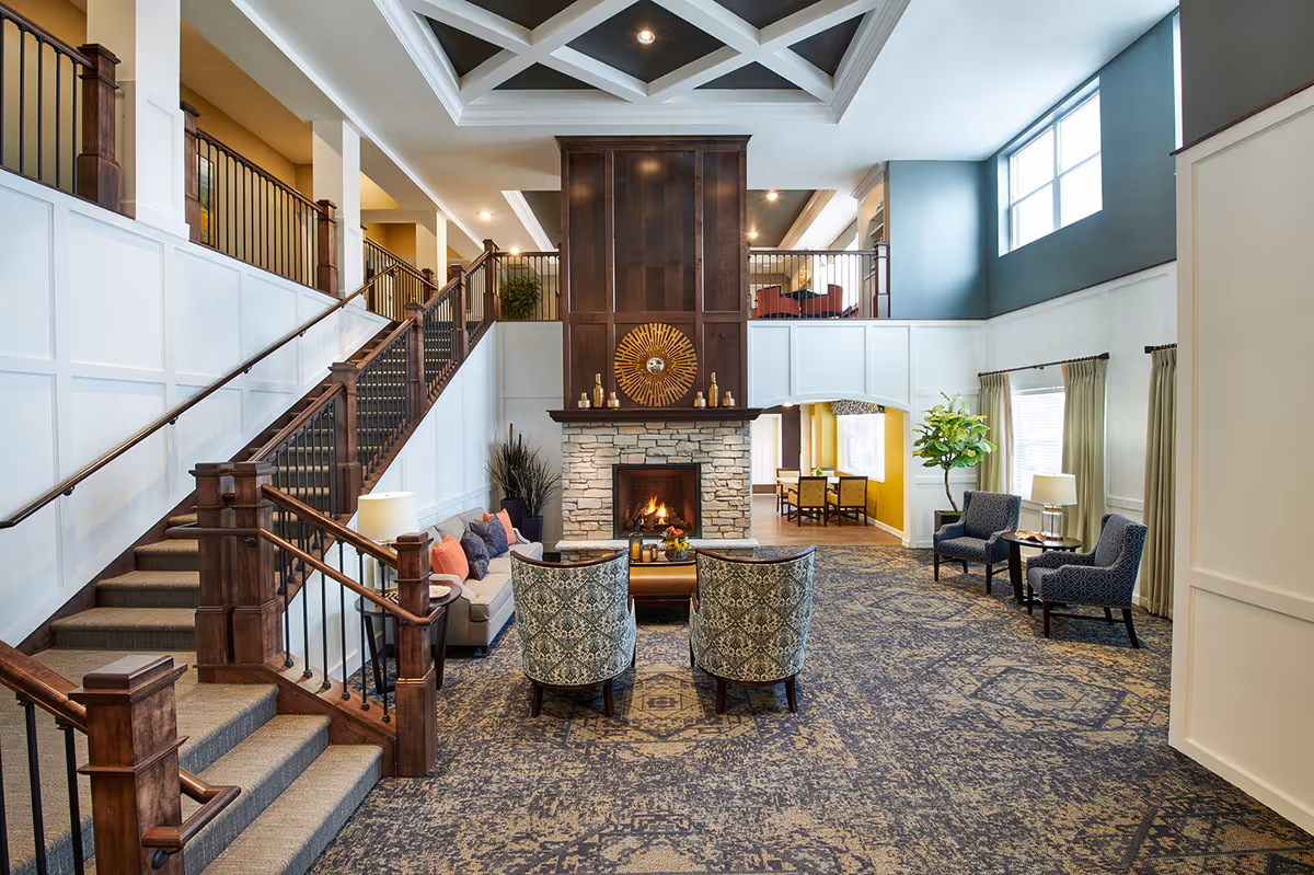 A spacious and well-lit living room area in a senior living facility featuring a stone fireplace with a wooden mantel and decorative sunburst mirror above it. The room has a high ceiling with a geometric design, large windows allowing natural light, and a patterned carpet. There are two patterned armchairs facing the fireplace, a beige sofa with colorful cushions, and additional seating with two blue armchairs near the windows. A staircase with wooden railings leads to an upper level with a balcony overlooking the room. A small dining area is visible through an archway in the background.