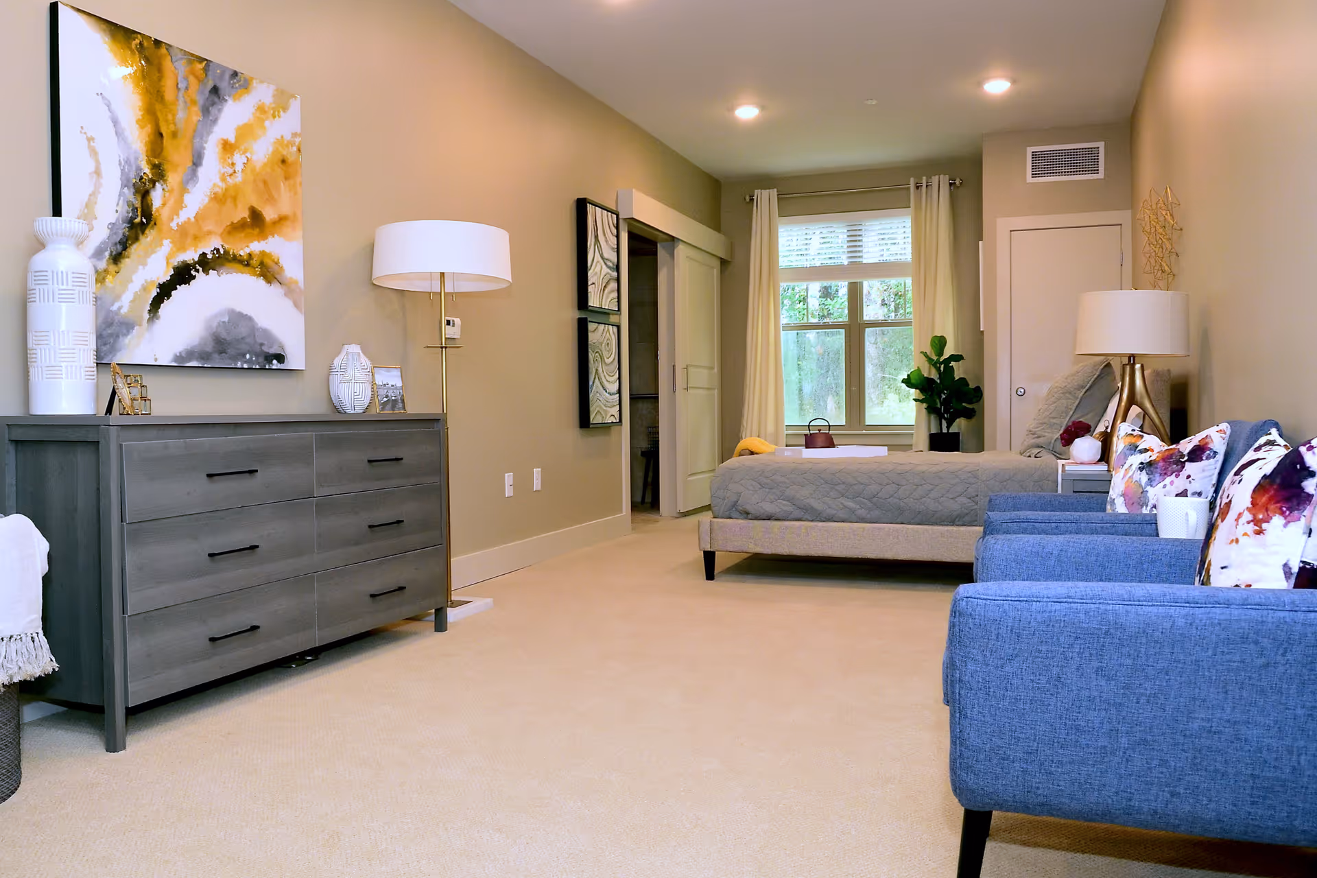 Sunlit bedroom with a bed, gray dresser, blue armchairs, lamps, and decorative artwork.