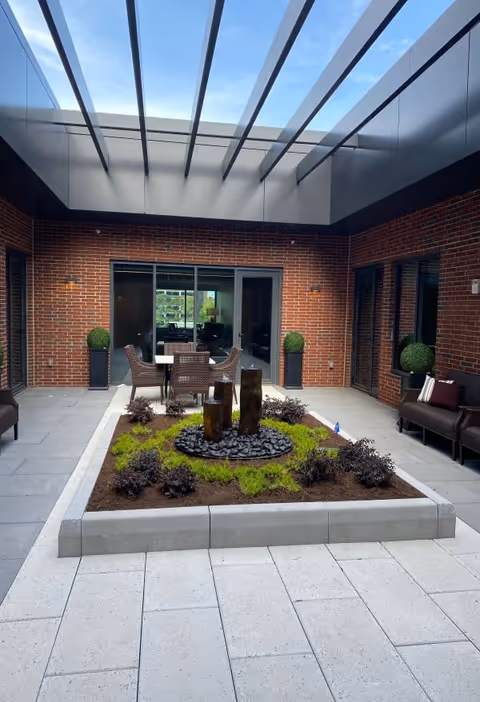 A modern indoor courtyard with a glass ceiling, featuring a central garden bed with plants and a small water fountain. Surrounding the garden are patio chairs and tables, with brick walls and large glass doors leading to interior rooms.