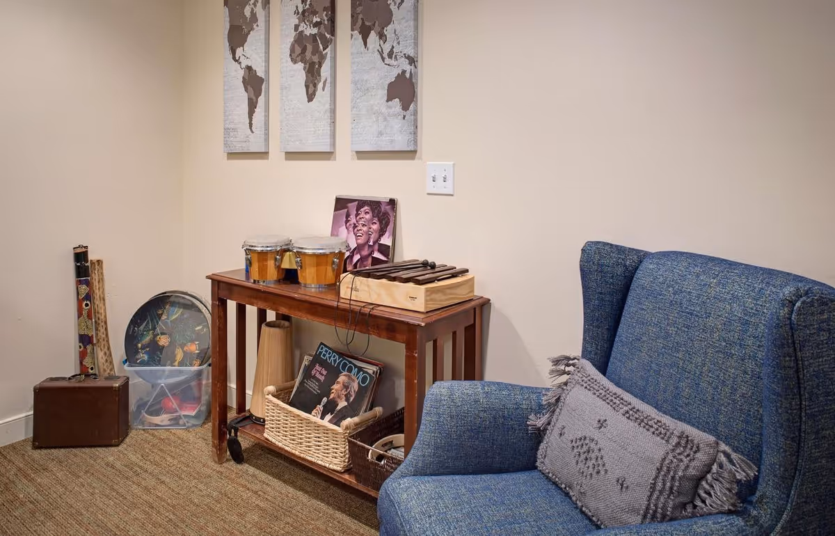 Corner of a cozy sitting area with a blue upholstered armchair, a wooden side table holding bongos, records and percussion instruments, and three world map prints on the wall.