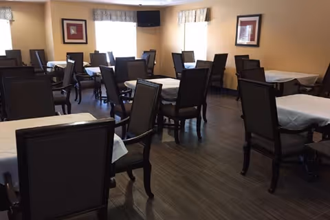 A dining room with multiple tables covered with white tablecloths and surrounded by dark wooden chairs. The room has light-colored walls, two windows with patterned valances, framed artwork on the walls, and a dark wood floor.
