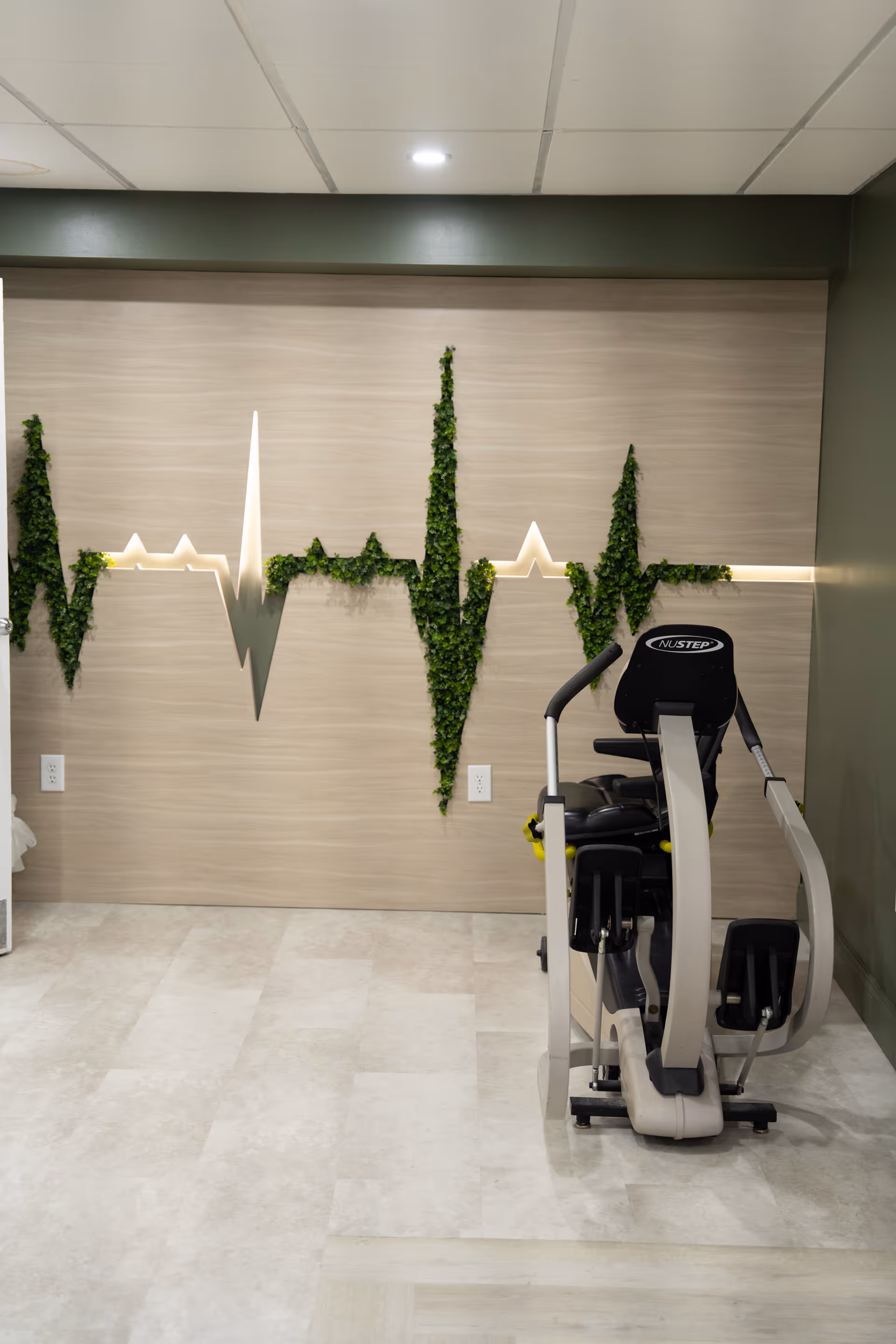Indoor exercise area with a NuStep recumbent cross trainer positioned on a light-colored tiled floor. The back wall features a decorative design resembling a heartbeat line made with green foliage and illuminated accents. The walls are painted in muted green and beige tones, and the ceiling has recessed lighting.