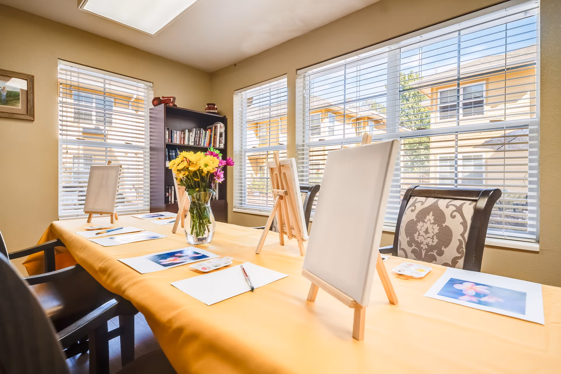 A bright room with large windows and blinds, featuring a table covered with a yellow tablecloth. On the table are blank canvases on easels, paint palettes, pens, and printed photos of flowers, suggesting an art activity setup. A vase with yellow and pink flowers is placed in the center of the table. In the background, there is a bookshelf filled with books and decorative items.