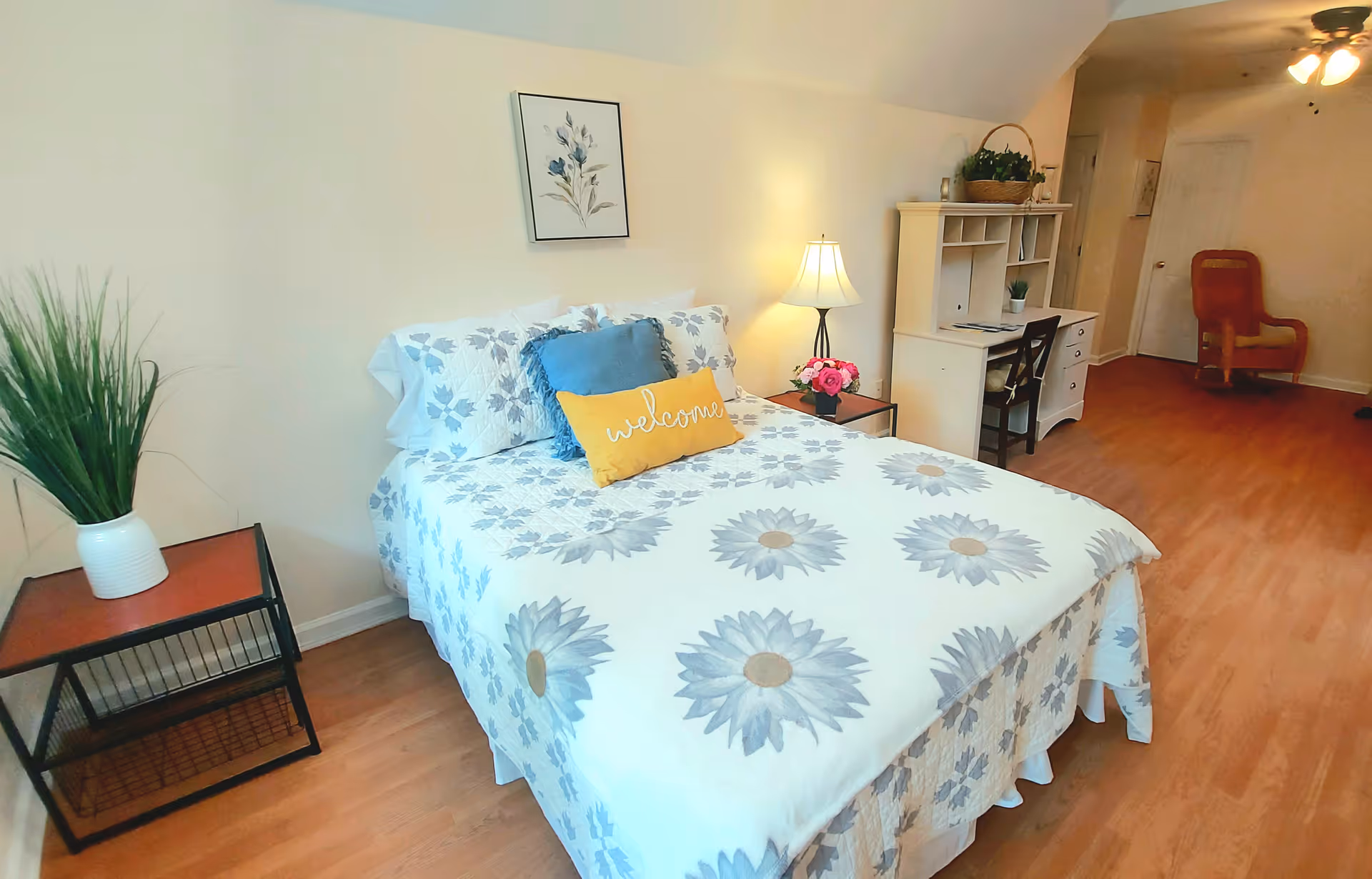 Cozy furnished bedroom featuring a double bed with a floral duvet and a 'welcome' pillow, nightstand, desk and wood flooring.