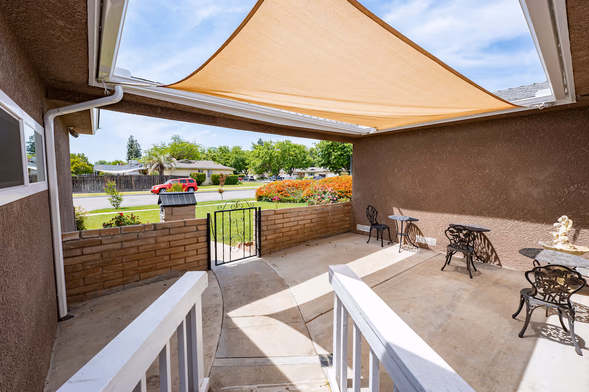 A covered outdoor patio area with a beige shade sail overhead, brown stucco walls, and a low brick wall with a black metal gate. The patio has several black metal chairs and small tables. Beyond the patio is a green lawn, bushes with flowers, and a street with houses and trees in the background under a partly cloudy sky.