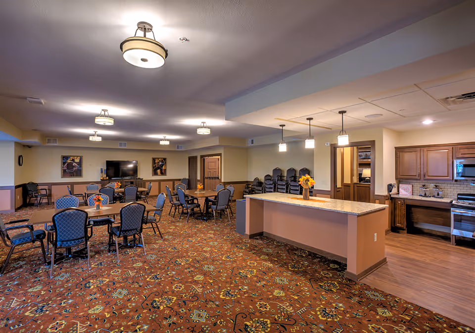A spacious senior living facility common area with multiple tables and chairs arranged for dining or socializing. The room features a patterned carpet, a kitchen area with wooden cabinets and a stove, a large island with pendant lights above, and a television mounted on the far wall. The walls are painted in neutral tones, and there are framed pictures on the walls.