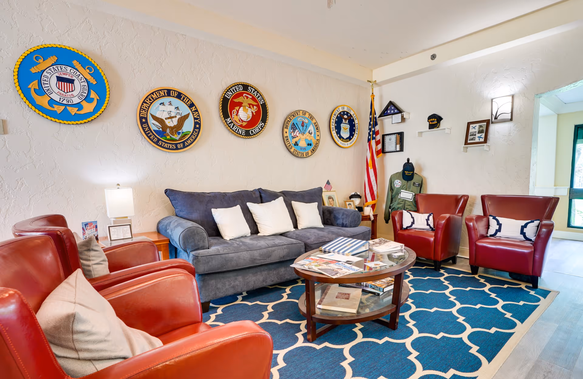 A cozy living room with a blue sofa and three white pillows, two red leather armchairs with beige pillows, and a round wooden coffee table with books and magazines. The walls are decorated with military emblems and memorabilia, including an American flag, a folded flag in a display case, and framed pictures. The room has a blue patterned rug and a side table with a lamp.