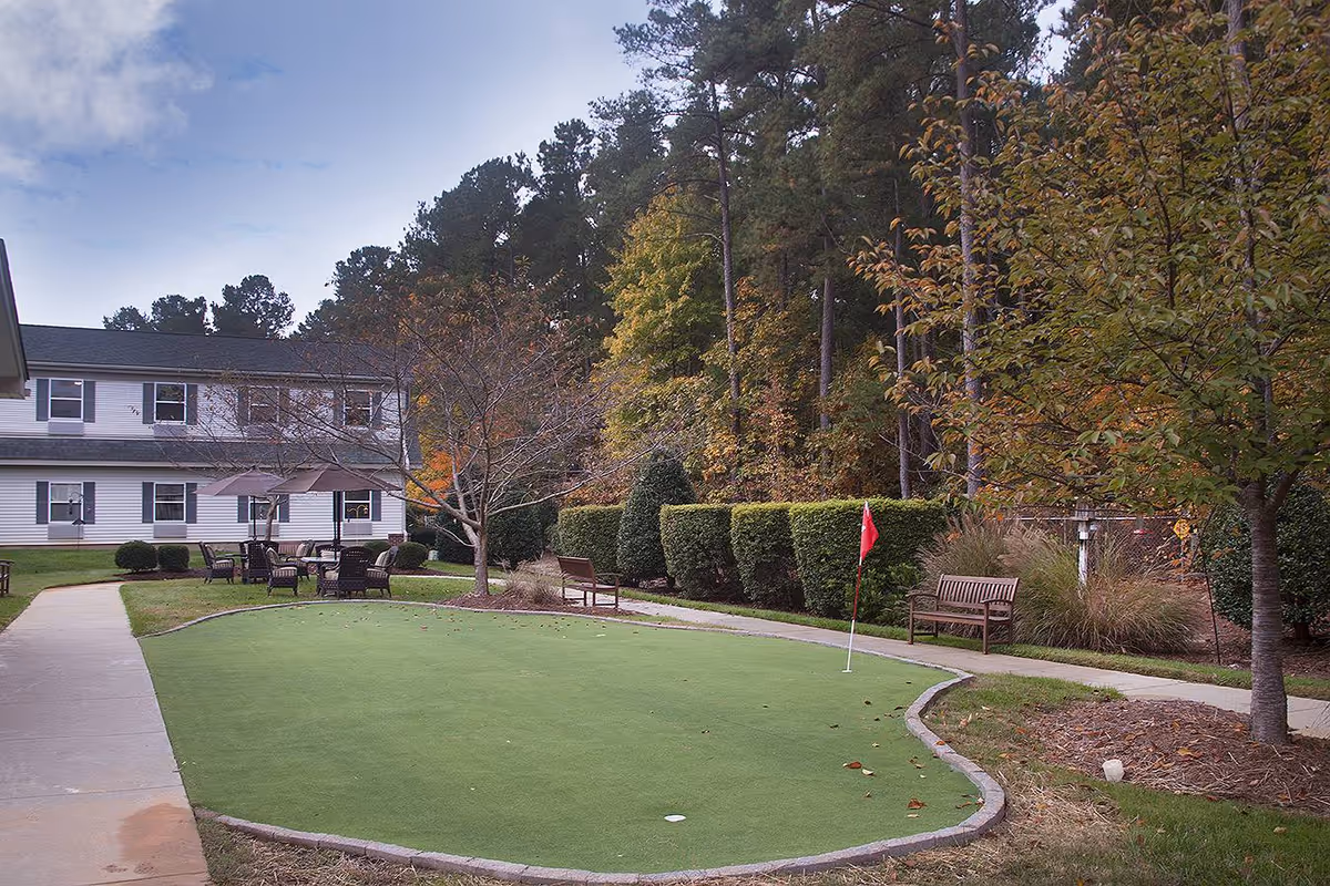 Outdoor area at Sunrise at North Hills featuring a putting green with a red flag, surrounded by benches, chairs, and tables. The area is bordered by trees and shrubs with a two-story building visible in the background under a partly cloudy sky.