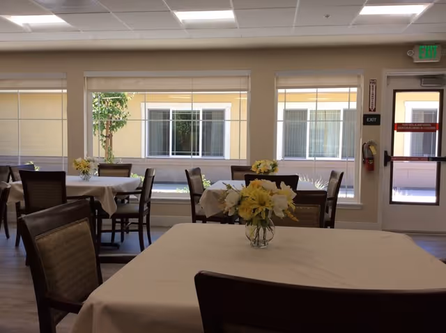 A dining room in a senior living facility with several tables covered in white tablecloths, each adorned with a vase of yellow and white flowers. The room has large windows letting in natural light and a door with an exit sign above it.