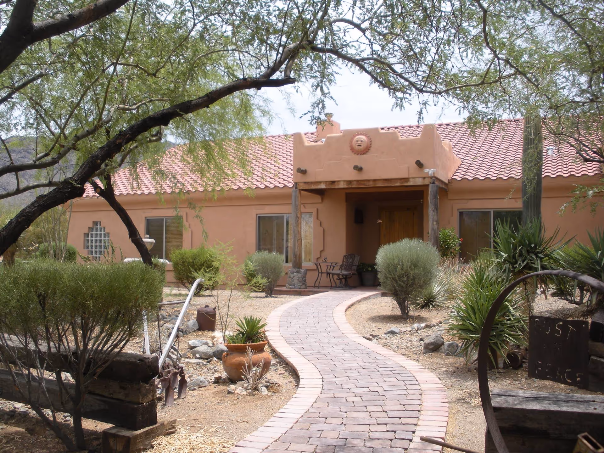A southwestern-style building with a curved brick pathway leading to a wooden front door. The building has a terracotta tile roof and adobe walls with decorative elements. The surrounding landscape features desert plants, shrubs, and trees, with a rustic bench and a sign that reads 'Trust in Peace'.