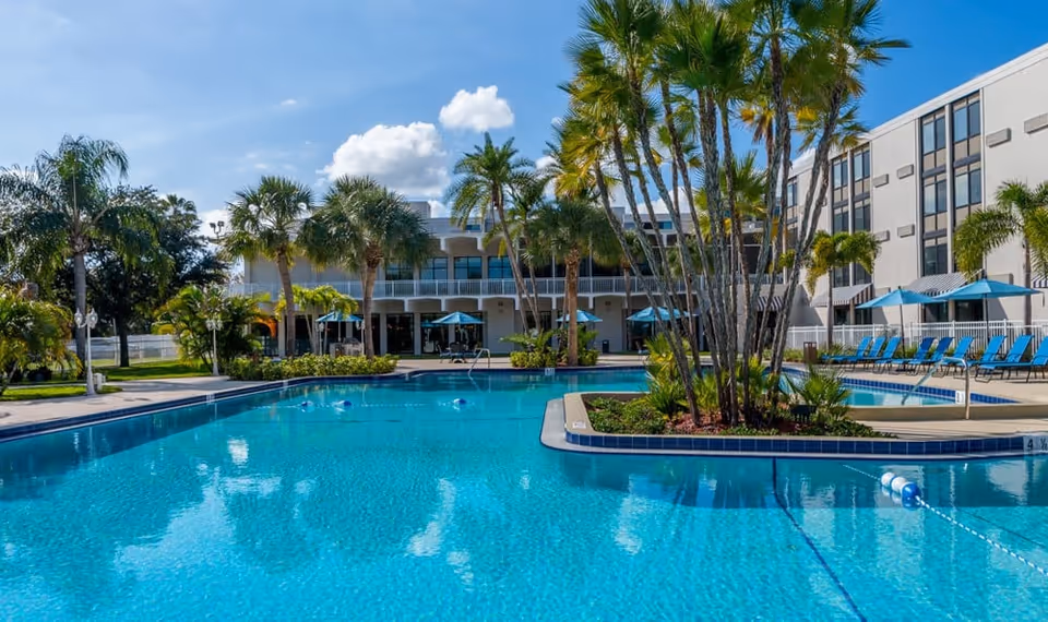 Outdoor swimming pool area at Volante Senior Living of St. Petersburg with clear blue water, palm trees, lounge chairs with blue umbrellas, and a multi-story building in the background under a partly cloudy sky.