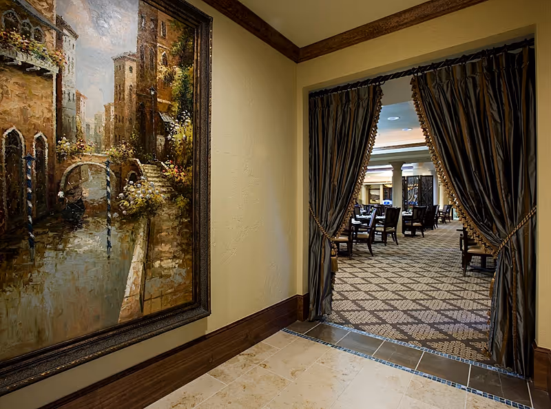 View through an open doorway with dark drapes tied back, leading into a carpeted dining area with multiple tables and chairs. On the left wall outside the doorway, there is a large framed painting depicting a canal scene with buildings and a bridge.