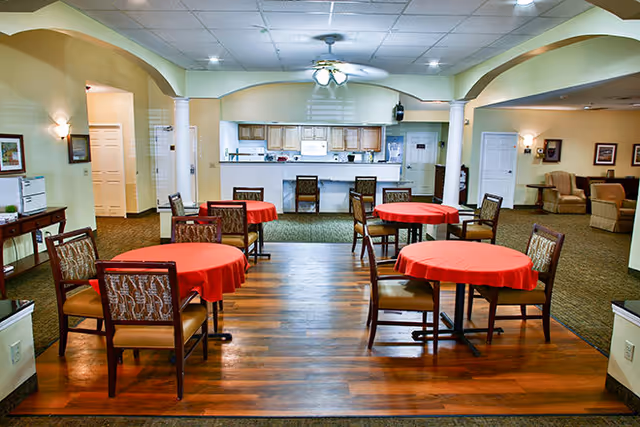 Interior view of a senior living facility dining area with four round tables covered with red tablecloths, each surrounded by four chairs. The room has a wooden floor section in the foreground and carpeted area in the background. There is a ceiling fan with lights, a kitchen counter with bar stools, and a seating area with armchairs in the back right corner.