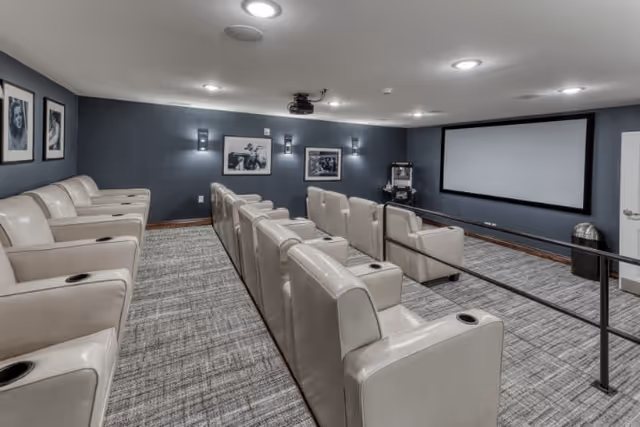 A small movie theater room with three rows of beige leather recliner chairs facing a large blank projection screen. The walls are painted dark gray and decorated with black and white framed photographs. The floor is carpeted in a patterned gray design, and there is a popcorn machine in the corner near the screen.