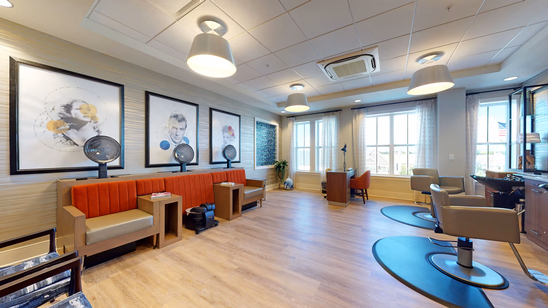 A bright and spacious salon area in an assisted living facility with large windows letting in natural light. The room features salon chairs with mirrors, hair dryers mounted on a red cushioned bench along the wall, modern ceiling lights, and framed black-and-white artwork on the walls.