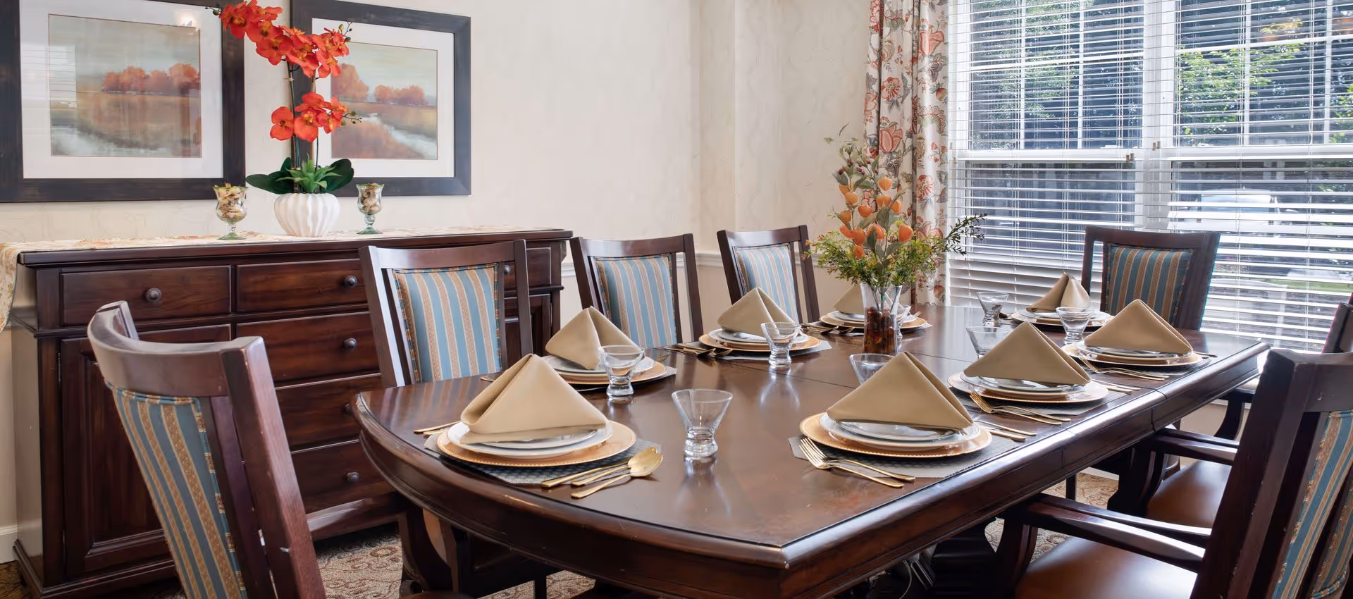 A formal dining room with a wooden table set for eight, folded napkins, chairs, a sideboard with decor, and a large window with blinds.