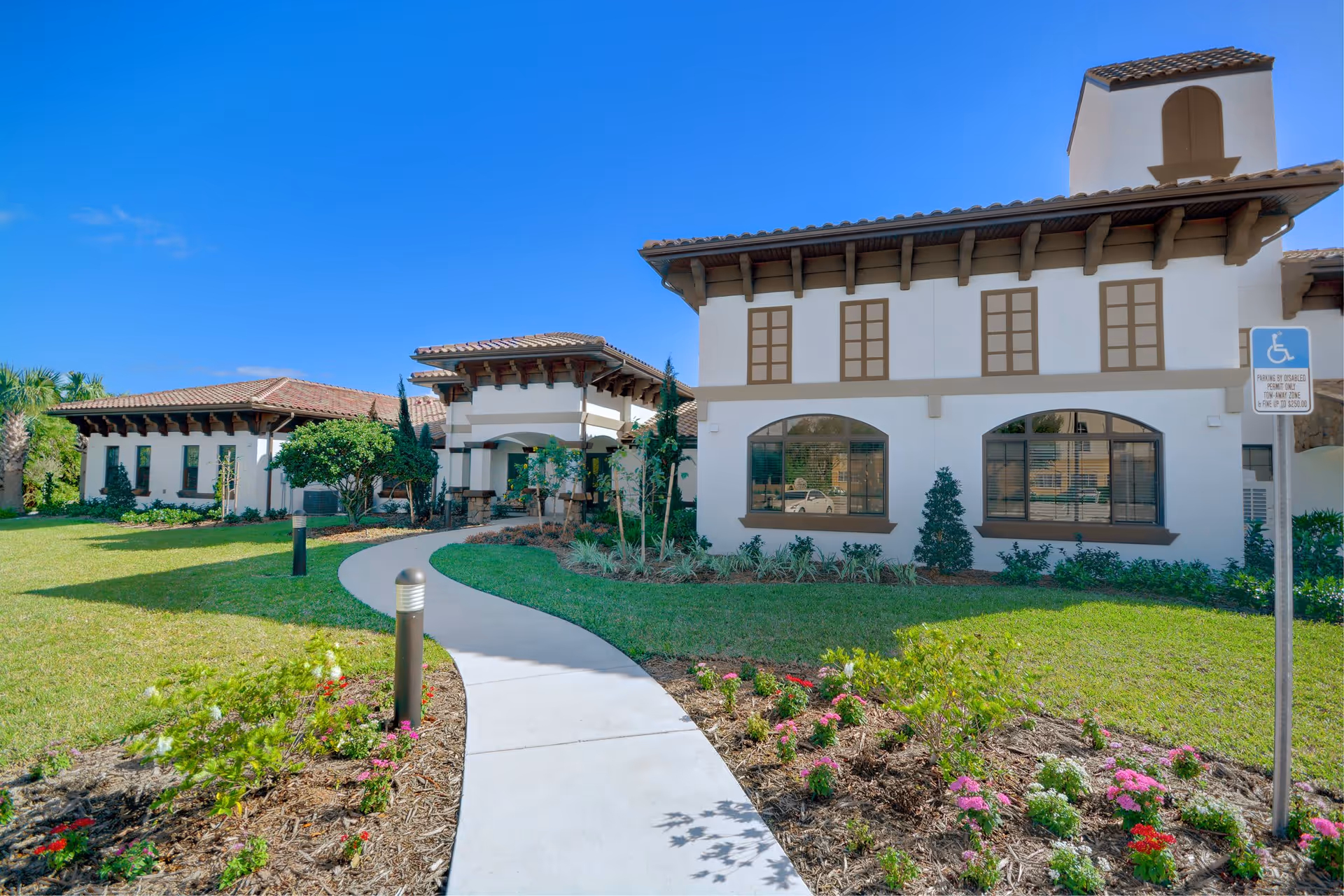 Exterior view of Tuscan Gardens Of Venetia Bay facility with a curved concrete walkway leading to the entrance. The building has a Mediterranean architectural style with white walls, brown trim, and a tiled roof. The landscaped garden features green grass, small shrubs, and colorful flowers under a clear blue sky.