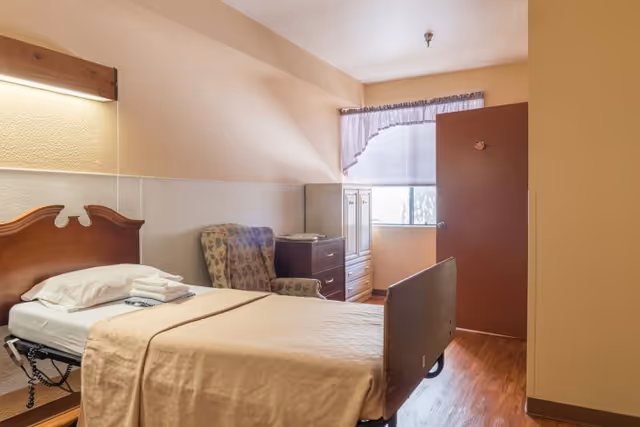 Single-occupancy senior living bedroom with a bed, armchair, dresser, and a window with a valance.