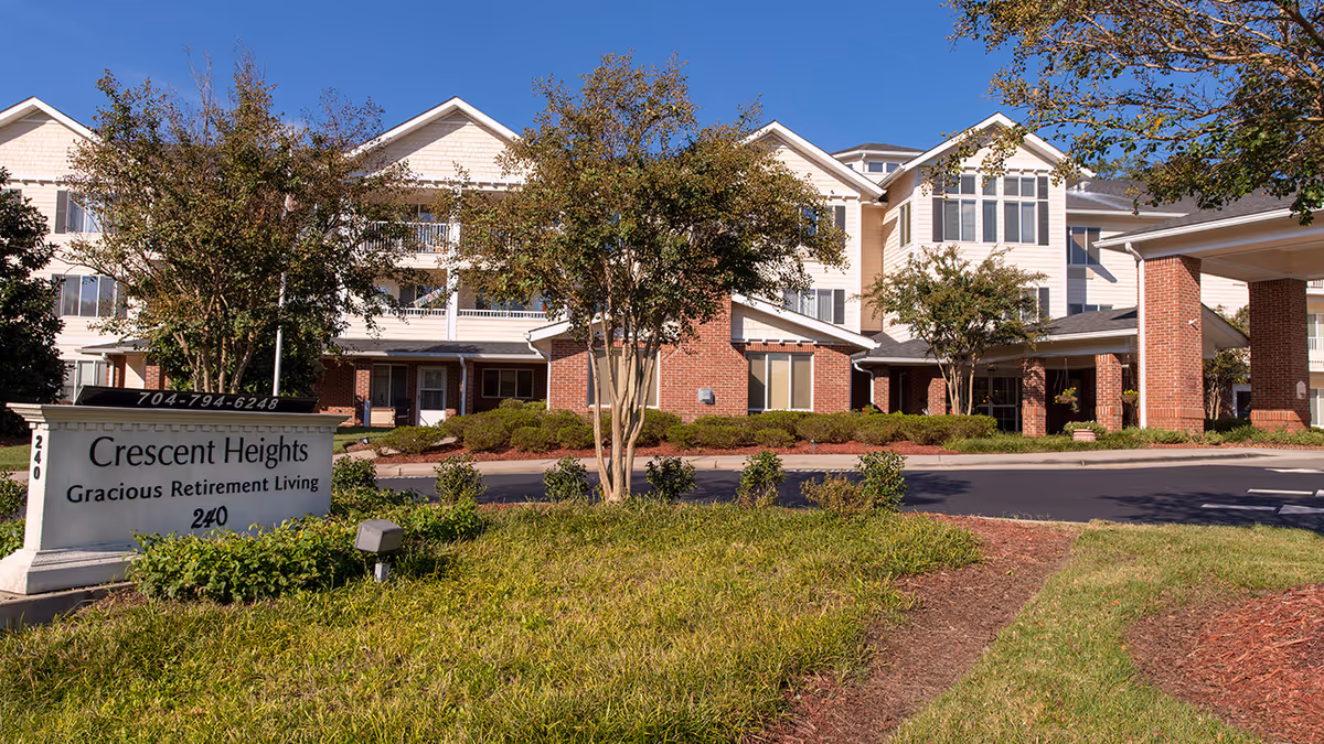 Exterior view of Crescent Heights retirement living facility showing a multi-story building with white siding and brick accents, surrounded by trees and landscaping under a clear blue sky. A sign in front reads 'Crescent Heights Gracious Retirement Living 240' with a phone number on top.
