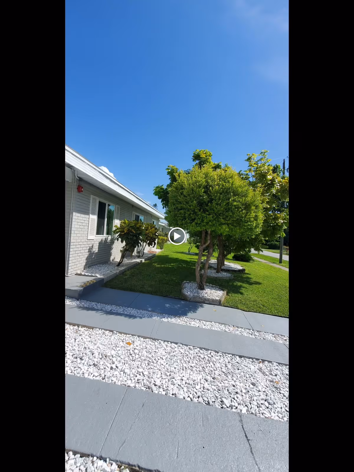 Exterior view of a single-story building with light gray brick walls and white window shutters. In front of the building, there is a landscaped area with neatly trimmed trees, green grass, and white decorative rocks bordered by gray concrete pathways. The sky is clear and blue.