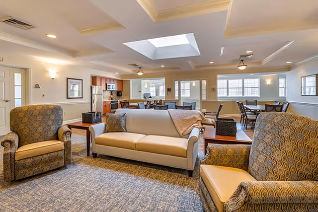 A spacious and well-lit common area in a senior living facility featuring a beige sofa with a throw blanket and two patterned armchairs arranged around a coffee table. In the background, there is a kitchen area with wooden cabinets, a refrigerator, and a microwave, as well as several dining tables and chairs near large windows allowing natural light to fill the room. The ceiling has a skylight and recessed lighting.
