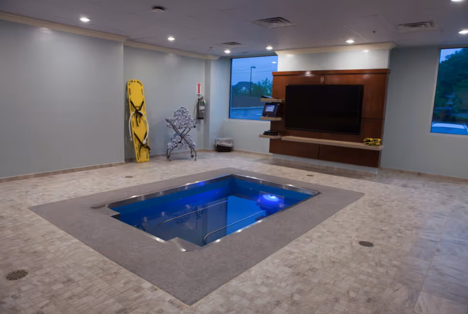 Indoor small recessed therapy pool in a tiled room with a wall-mounted TV, windows, and safety equipment against the wall.