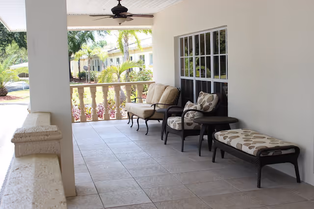 Covered outdoor patio area with cushioned wicker furniture including a loveseat, armchair, small round table, and a cushioned bench. The patio has tiled flooring, a ceiling fan, and overlooks a garden with palm trees and other greenery.