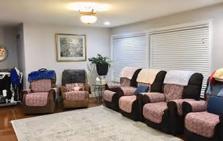 A cozy living room with multiple recliner chairs covered with pink and black slipcovers, a large window with white blinds, a light-colored area rug on wooden flooring, a framed painting on the wall, and a small side table with a plant.