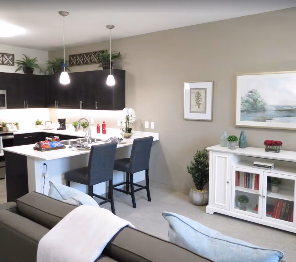 Modern kitchen and living area with a white island countertop, two dark upholstered bar stools, dark cabinetry, pendant lights, and decorative plants. A white cabinet with books and decorative items is against the wall, along with framed artwork.