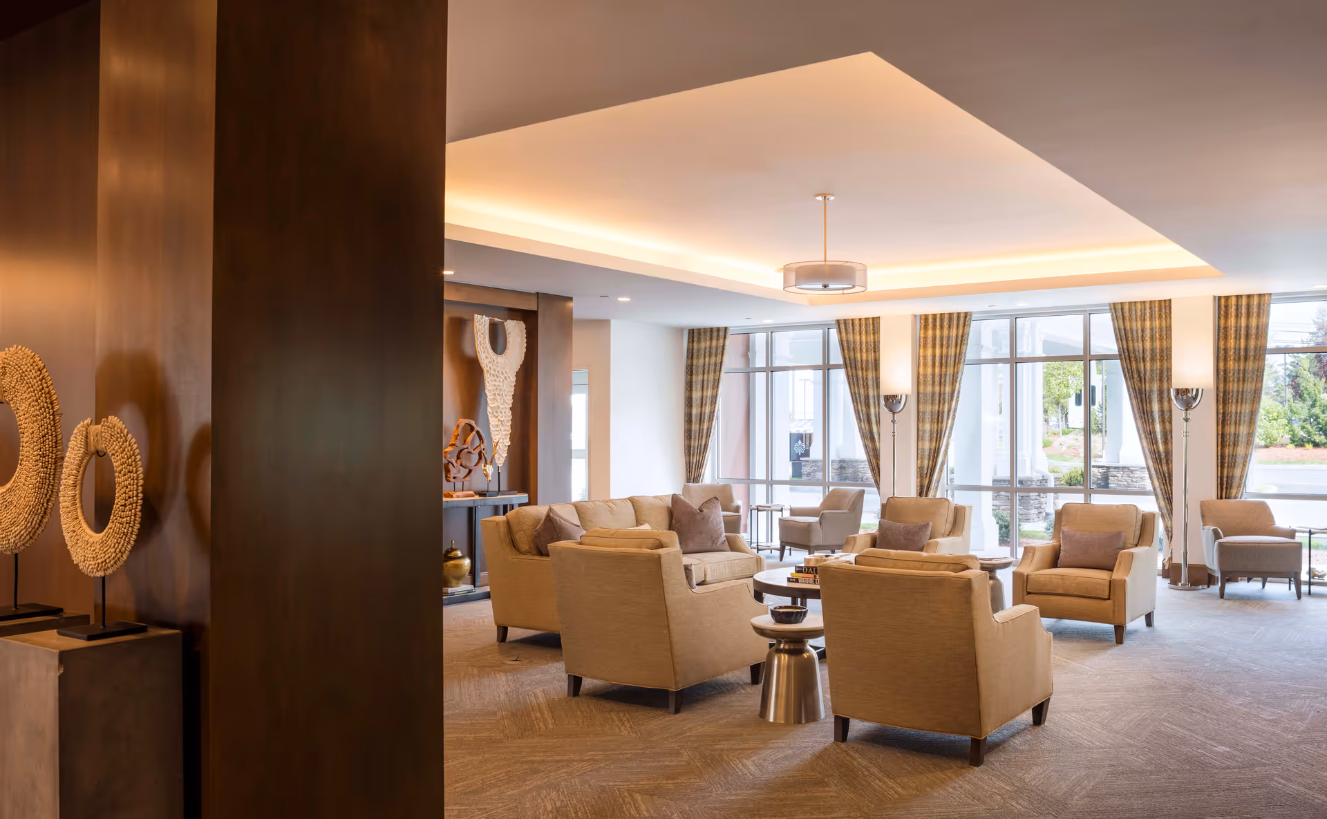 A spacious and well-lit living room area with beige armchairs and sofas arranged around small tables. Large windows with plaid curtains allow natural light to fill the room. Decorative art pieces are displayed on a dark wooden wall on the left side.