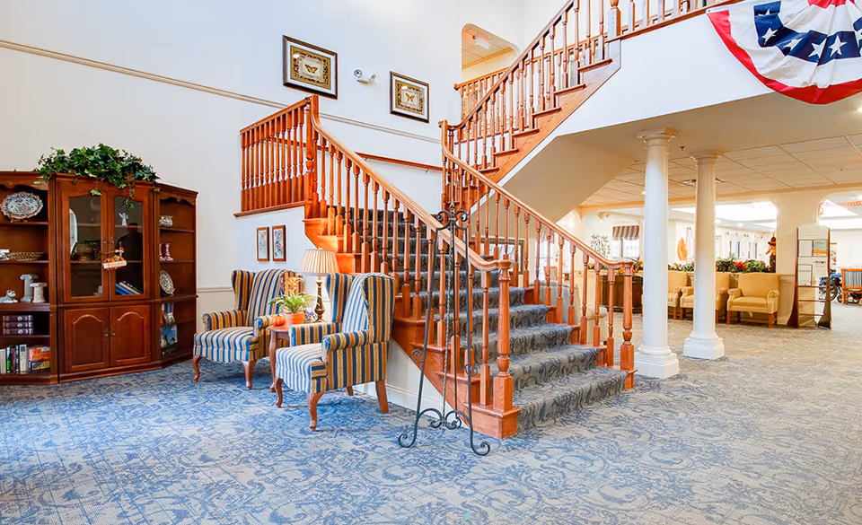 Bright community lobby with a central carpeted wooden staircase, two striped armchairs, a display cabinet, and seating under a balcony.