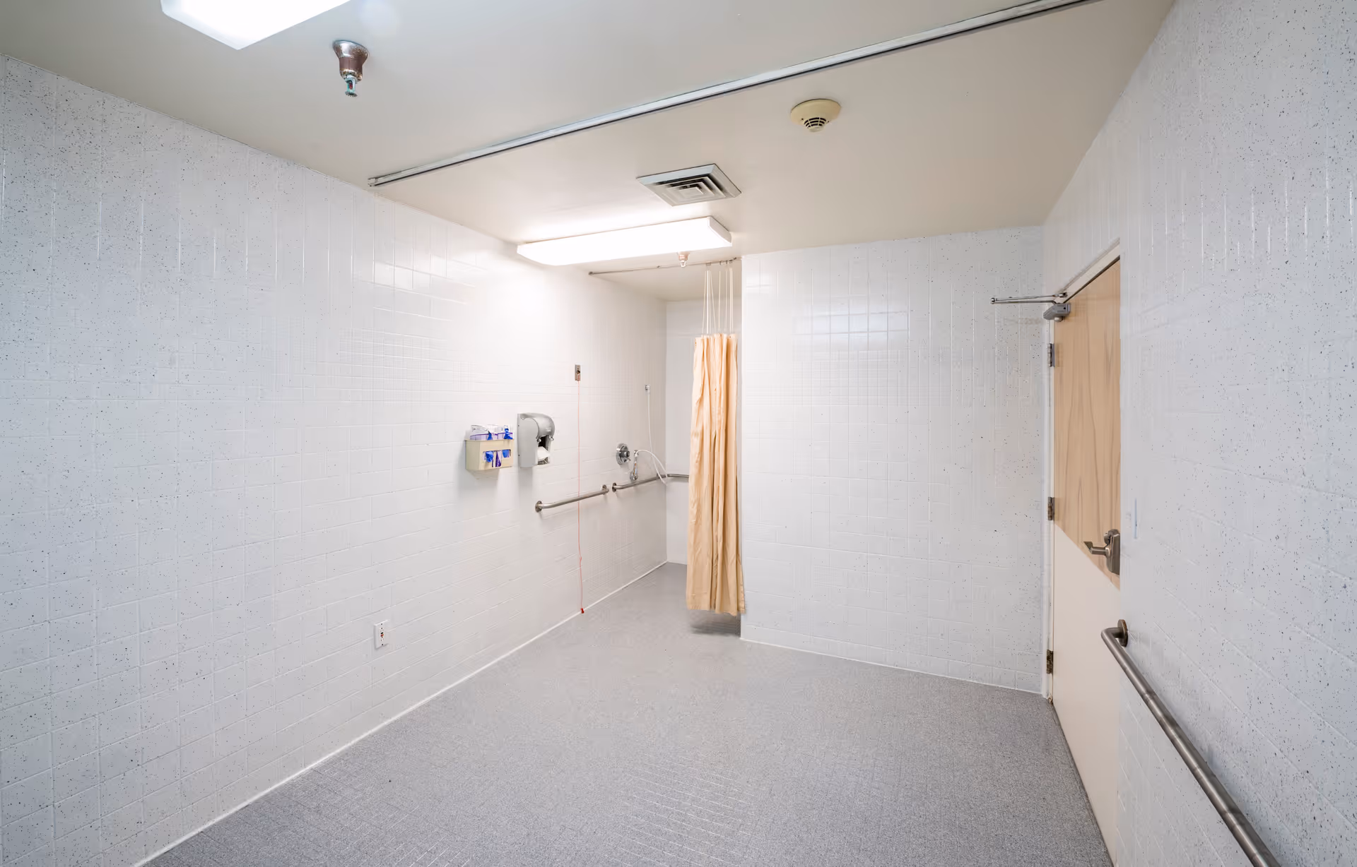 A clean, empty shower room with white tiled walls and a gray floor. There is a beige shower curtain hanging from the ceiling, a wall-mounted soap dispenser, a handrail, and a door with a handle on the right side. The ceiling has a fluorescent light fixture and ventilation.