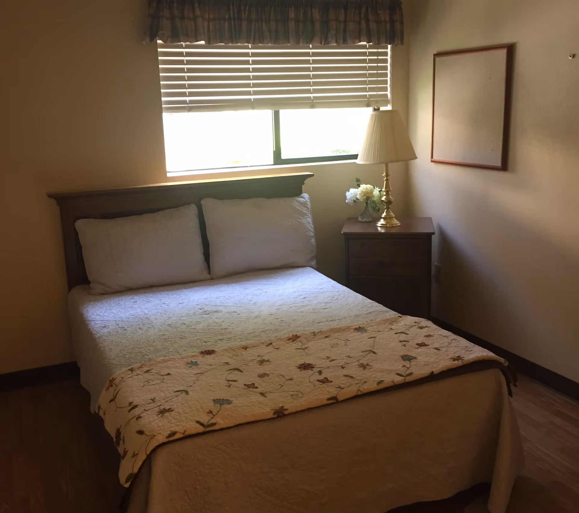 A bedroom with a neatly made bed featuring white bedding and a floral quilt at the foot. There are two white pillows against a wooden headboard. Next to the bed is a wooden nightstand with a brass lamp and a small vase of white flowers. A window with closed blinds and a valance is above the headboard. A blank framed board hangs on the wall to the right.