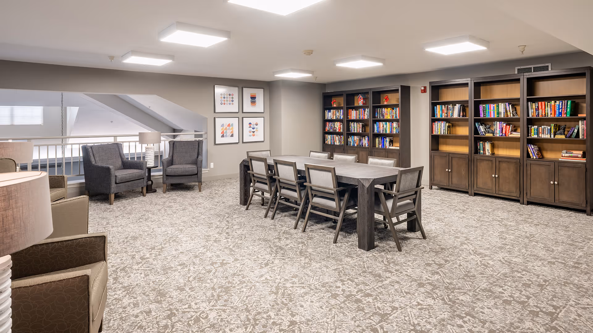 A spacious, well-lit common area with a large rectangular table surrounded by eight chairs in the center. Along the back wall are several dark wood bookshelves filled with books and decorative items. To the left, there are two gray armchairs with a small round table and a lamp between them. The floor is carpeted with a patterned design, and the walls are painted a light neutral color. Three framed abstract art pieces hang on the wall near the armchairs. The ceiling has recessed lighting panels.