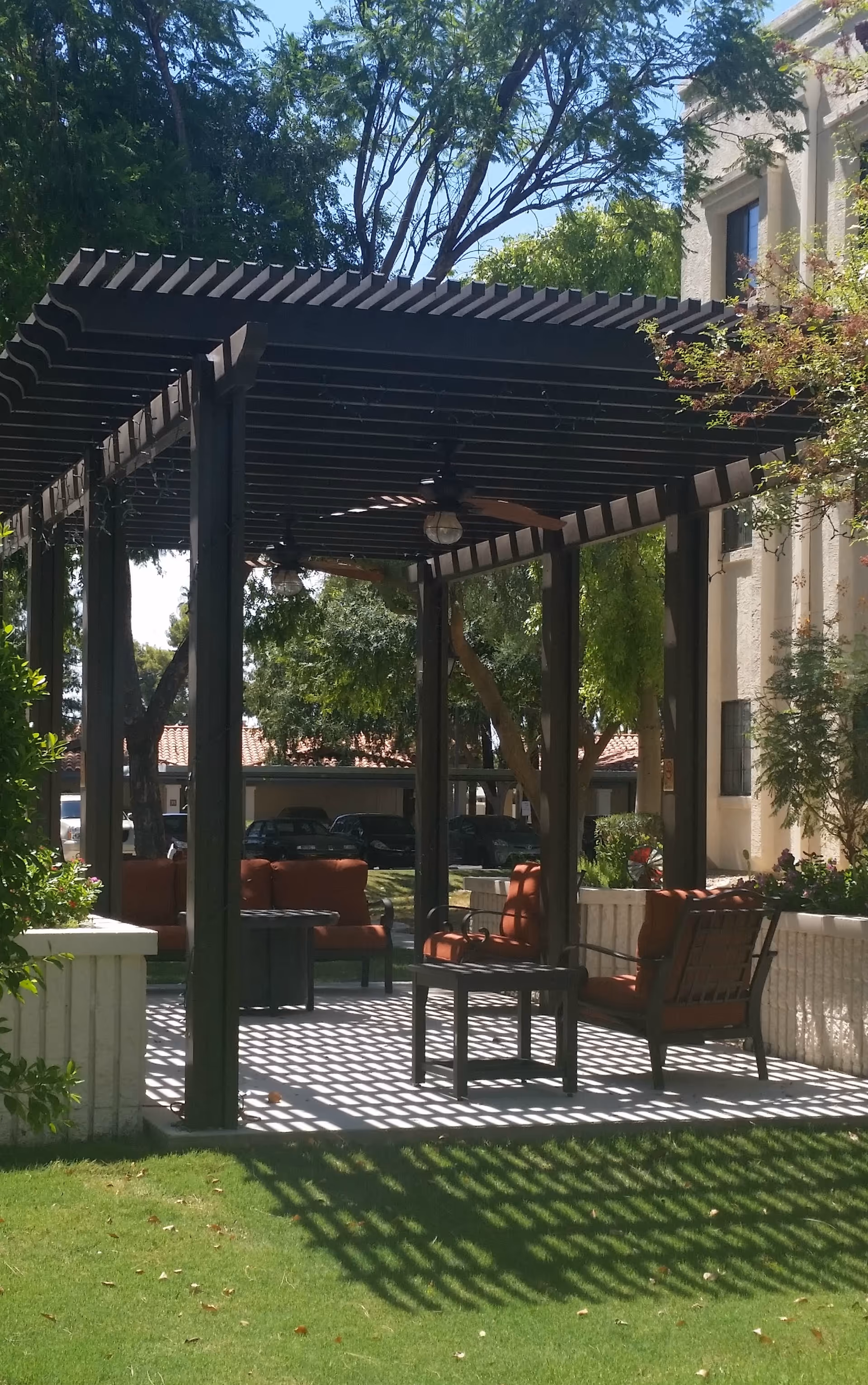 Outdoor seating area with cushioned chairs and tables under a wooden pergola casting shadows on the ground, surrounded by trees and greenery near a building.