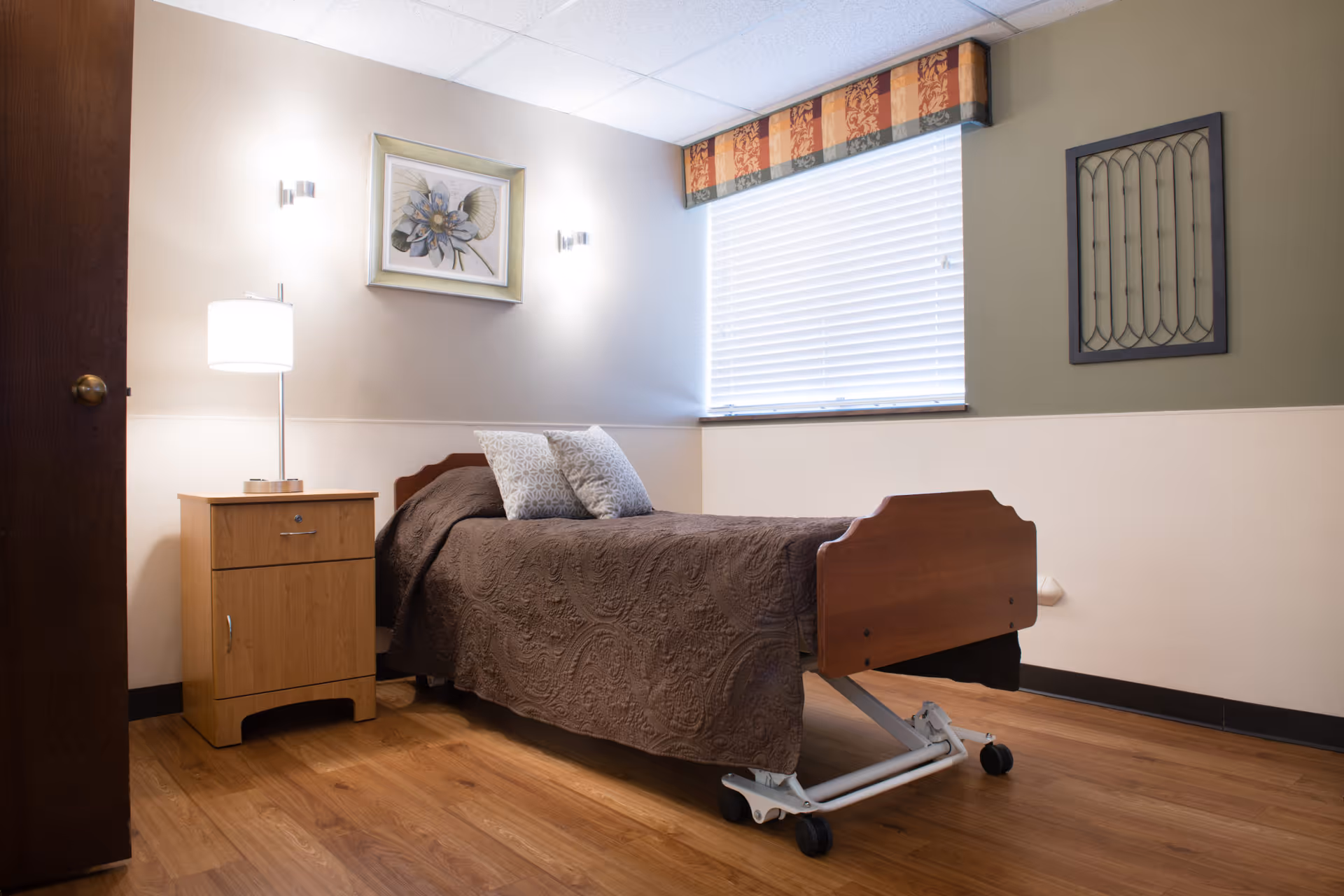 A simple senior bedroom with a single adjustable bed covered in a brown quilt, a wooden nightstand with a lamp, and a window with blinds.