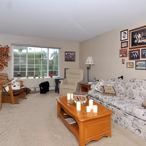 A cozy living room with a patterned sofa, a wooden coffee table with candles, an armchair, and a large window letting in natural light. The walls are decorated with framed pictures and artwork.