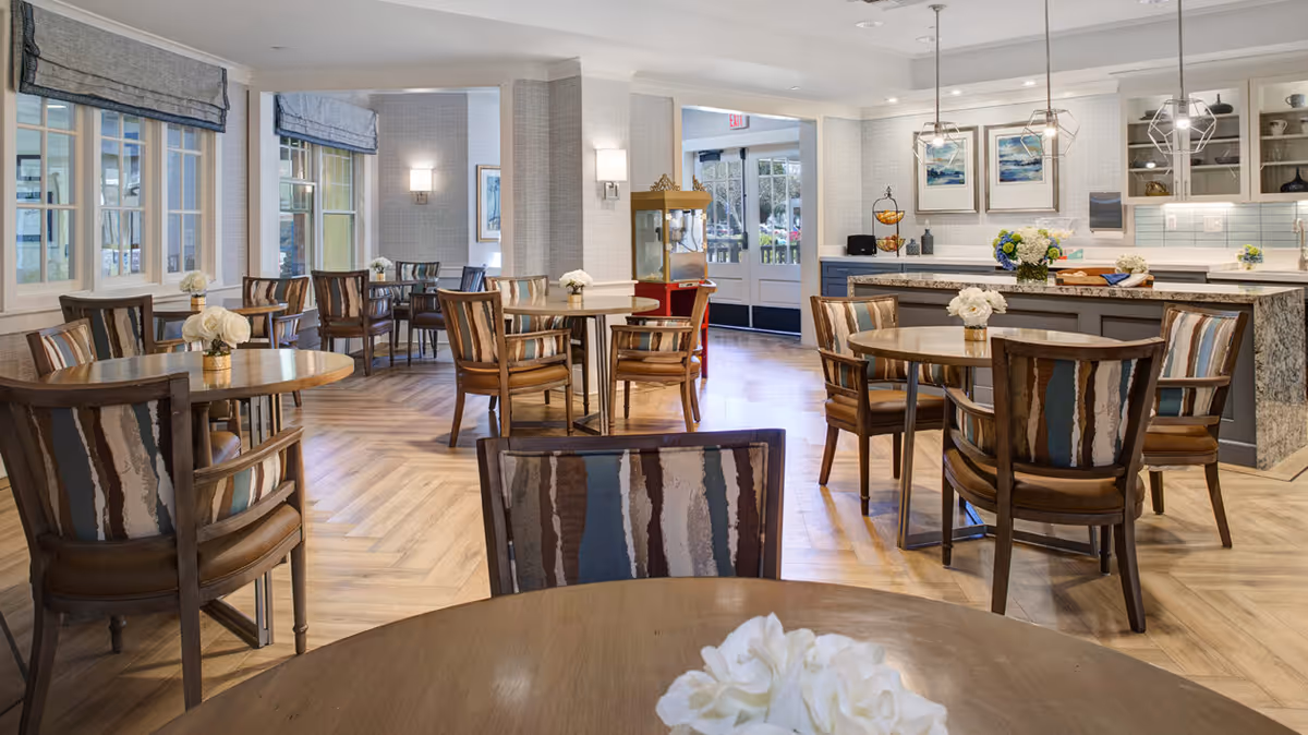 A bright and spacious dining area with multiple round wooden tables and cushioned chairs featuring striped upholstery. The room has large windows with blue roman shades, light wood herringbone flooring, and soft wall sconces. In the background, there is a kitchen area with a granite countertop island, pendant lights, and decorative flowers on the tables and counter.
