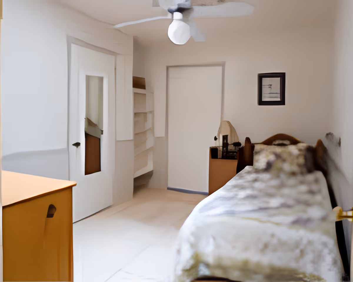 A narrow, minimally furnished bedroom with a single bed along the right wall, a dresser on the left, built-in shelving, and a ceiling fan.