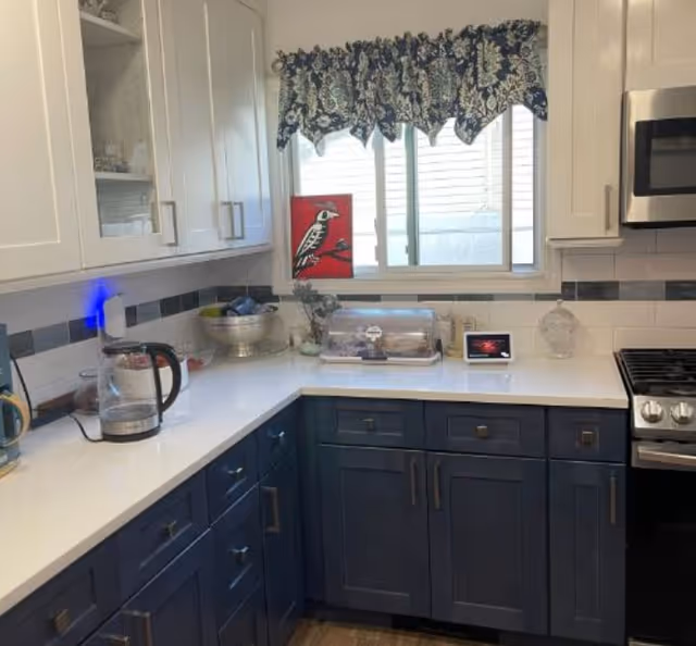 A modern kitchen corner with white upper cabinets and dark blue lower cabinets. The countertop is white with various items including a glass electric kettle, a silver bowl, a covered cake stand, and a small digital display. A window with a blue and white patterned valance is above the counter, and a stainless steel stove and microwave are visible on the right side.