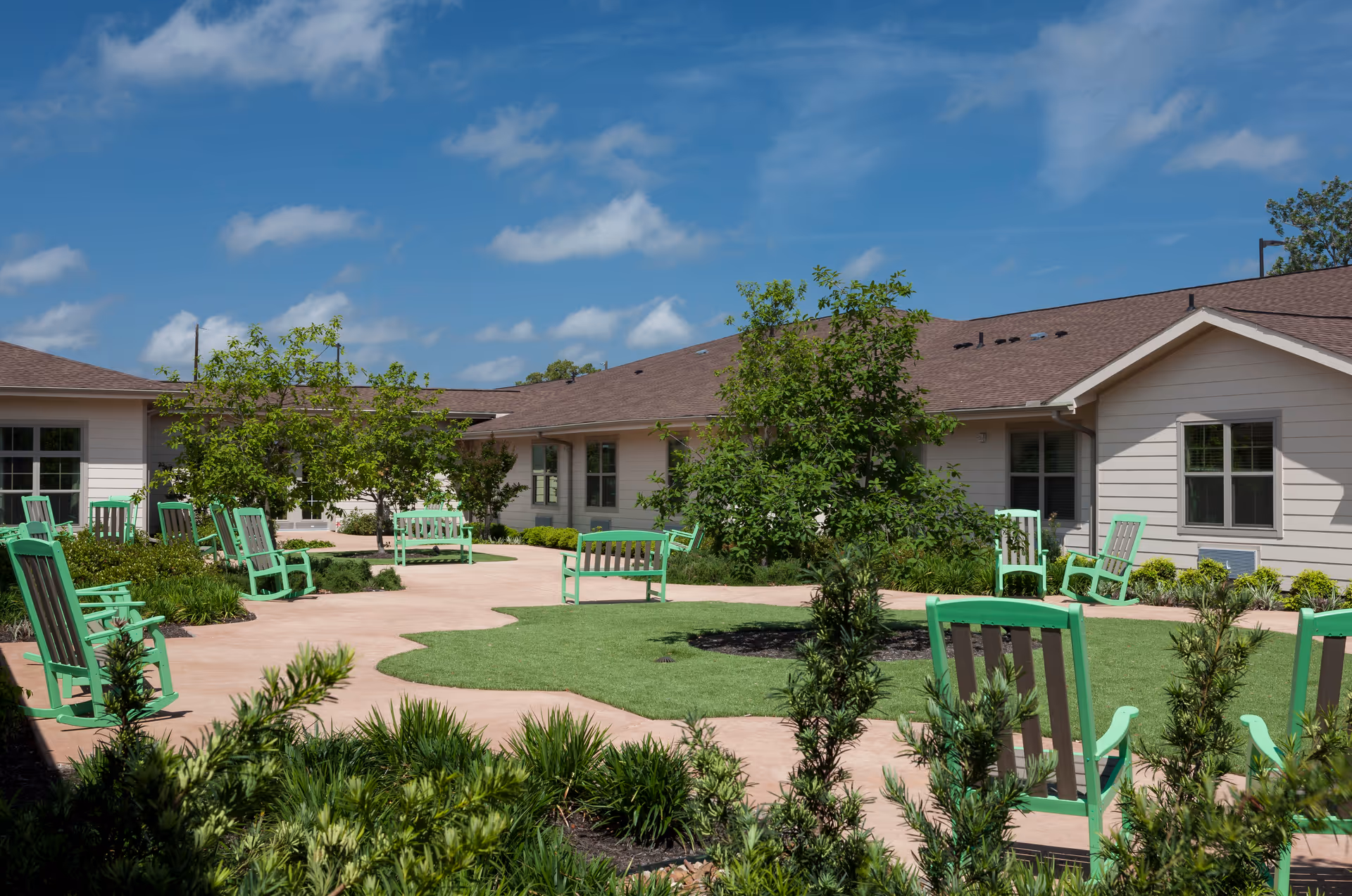 Outdoor courtyard area at Franklin Park Boerne featuring green rocking chairs and benches arranged on a paved walkway surrounding a grassy area with small trees and shrubs, under a blue sky with scattered clouds.