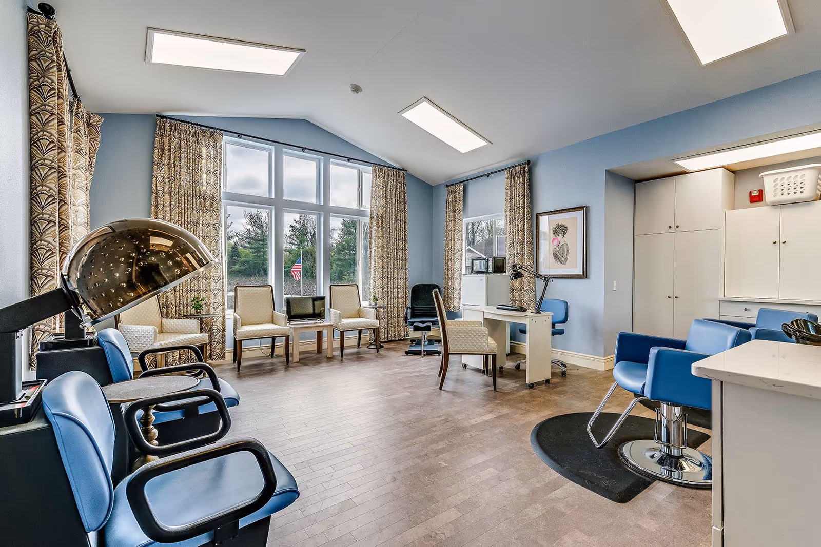 A bright and spacious room with large windows and patterned curtains letting in natural light. The room features several blue salon chairs, a hair dryer, a white desk with a chair, and additional seating with beige upholstered chairs. The walls are painted light blue and there are cabinets and a countertop on one side, suggesting a salon or grooming area within a senior living facility.