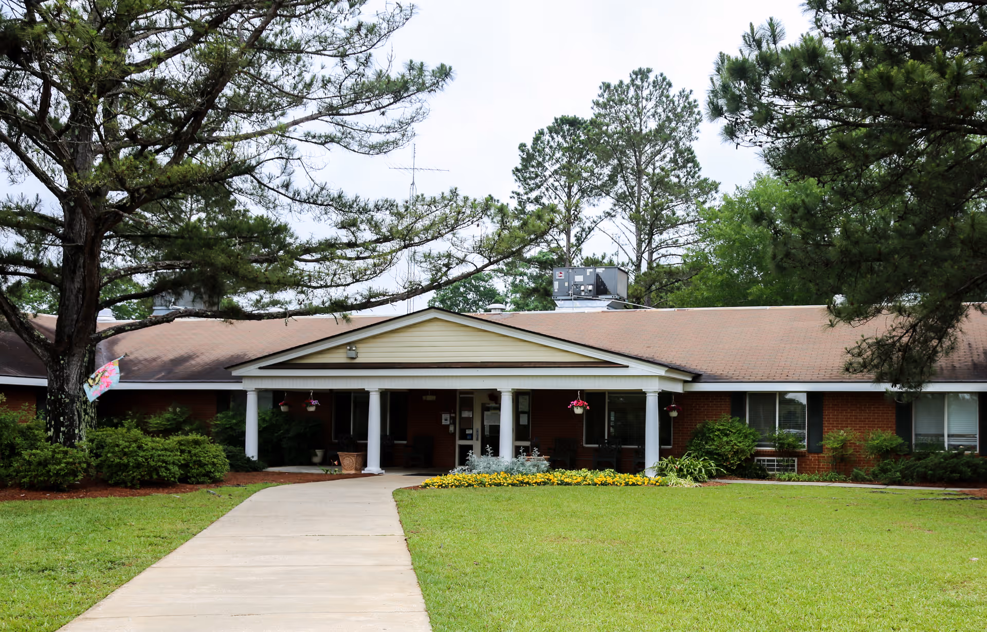 Front exterior view of a single-story brick building with a covered entrance supported by white columns. The building is surrounded by green grass, trees, and shrubs, with a concrete walkway leading up to the entrance. There are hanging flower pots under the covered entrance.